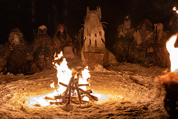 Antler Queen sits behind a fire surrounded by people in ritualistic outfits