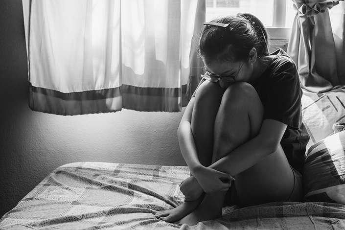 Young woman wearing shorts sitting on a bed with her legs pulled up under her chin looking scared or sad.