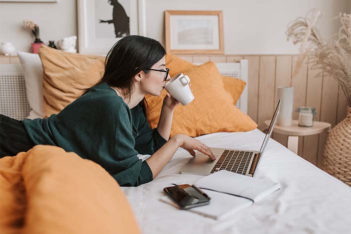 white woman with dark hair working on computer on bed