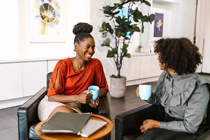 Two Black women talking with coffee mugs in their hands.