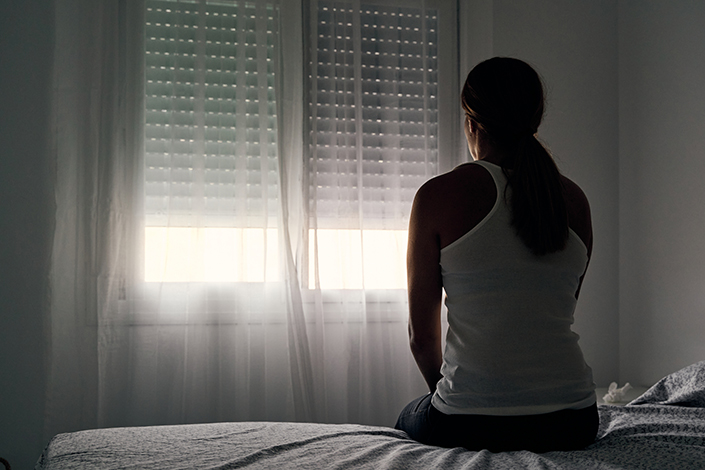 View of the back of a young woman wearing a white tank top sitting on a bed in front of partially closed blinds.