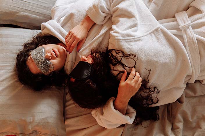 two women with dark hair embrace while wearing eye masks and laying in bed