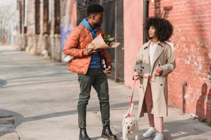 Two people in the streets stopped to talk, one holding flowers the other walking a dog