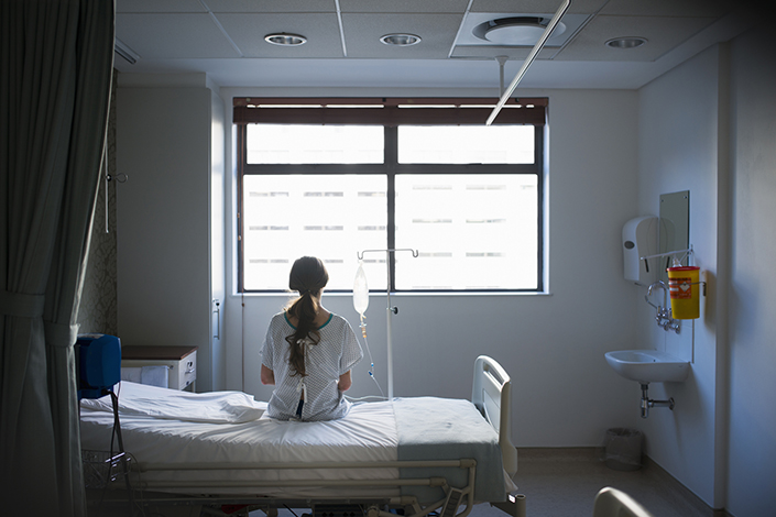 The back of a Hispanic woman sitting on a hospital bed next to an IV pole, looking out the window. 