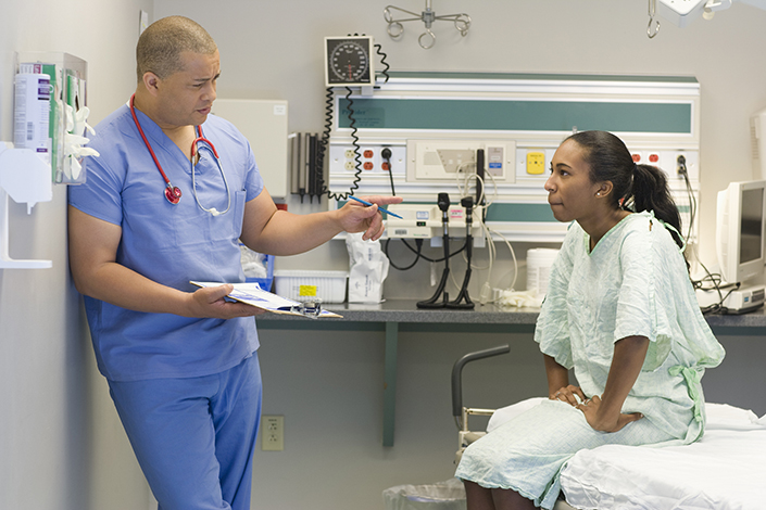 A young African American woman sitting on a hospital bed wearing a hospital gown listening to a male doctor, who is standing in front of her gesturing with his hands, explain something to her. 
