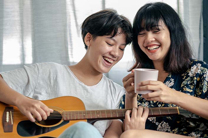 Two people laughing on a ouch, one with a guitar and one with a cup in hand