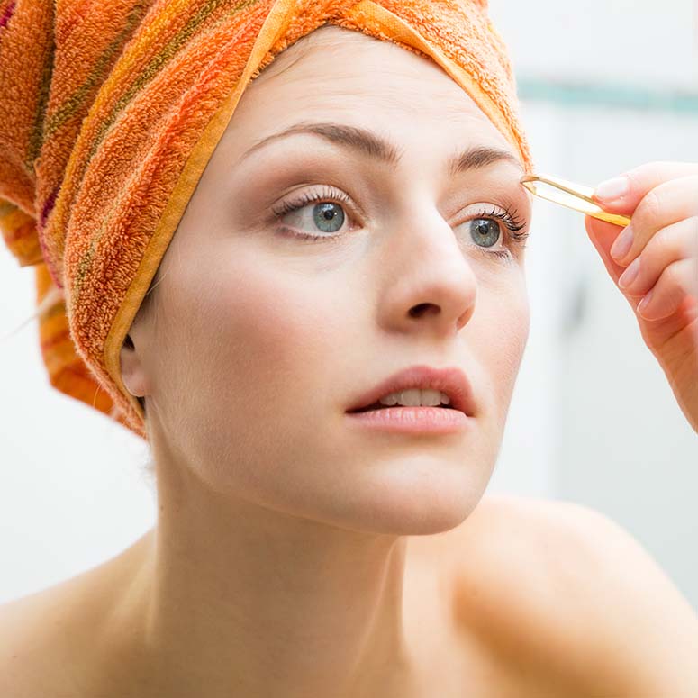 woman with her hair wrapped in an orange towel plucking her eyebrows with tweezers looking at her reflection in the mirror