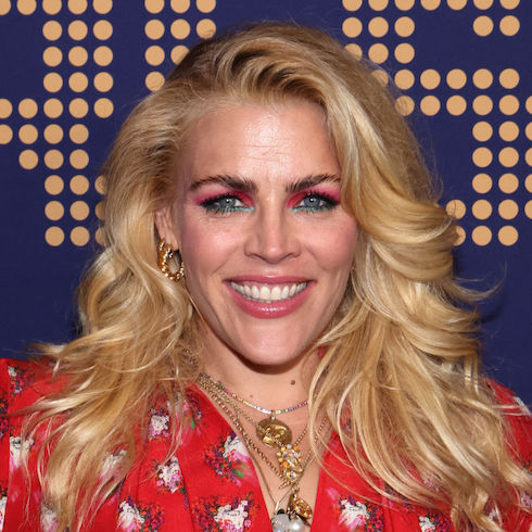Actress Busy Philipps wearing a red patterned top, hair down and feathered, smiling at the camera.