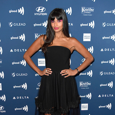 Photo of actress Jameela Jamil with her hands on her hips, standing in front of a blue and white background wearing a strapless black dress.