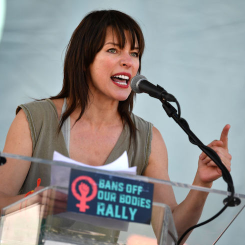 Actress Milla Jovovich passionately speaking into a microphone at a podium with a sign reading 'Bans off our bodies rally'.