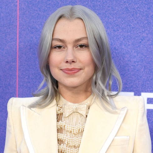 Singer Phoebe Bridgers wearing a cream-coloured blazer and smiling in front of a purple background.