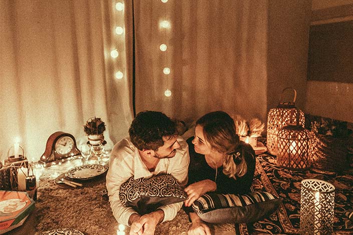 A smiling young couple laying on their stomachs on a pile of blankets and pillows, surrounded by twinkle lights