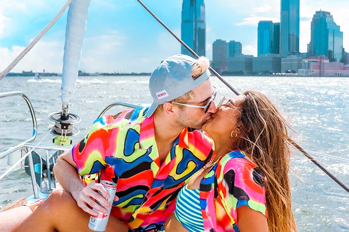 Young man and woman, both wearing brightly coloured clothes, sharing a kiss while on a boat with a city skyline in the background
