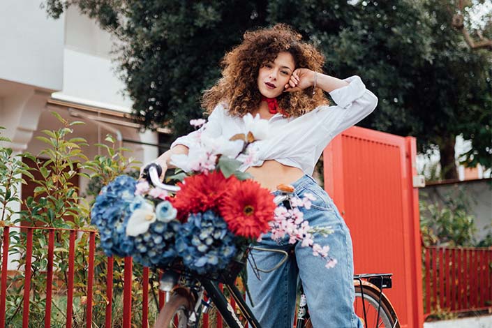 A young woman with curly hair stands by a bike with a variety of colourful flowers at the front
