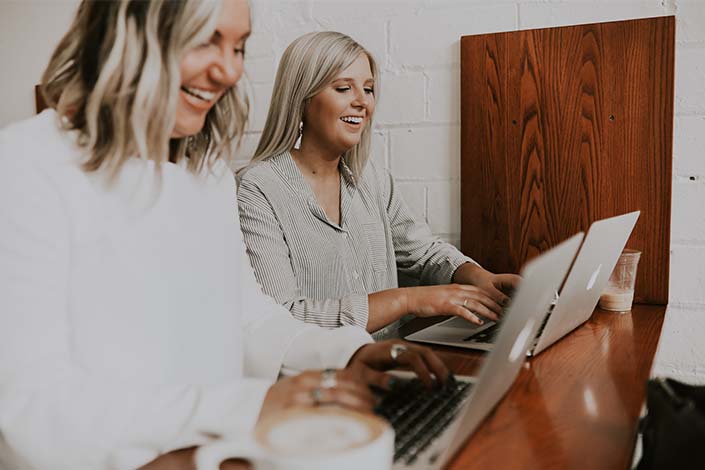 Two blonde women sitting side by side while working on laptop computers and smiling
