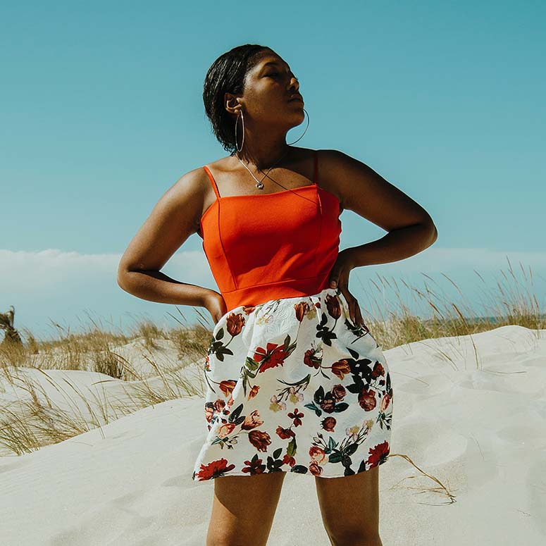Young Black woman wearing a bright red-and-white outfit while standing on a sandy beach on a blue-sky day