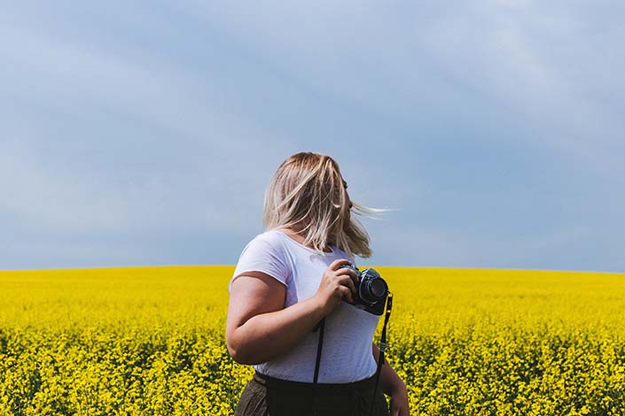 A blonde woman stands in a yellow field while holding a camera