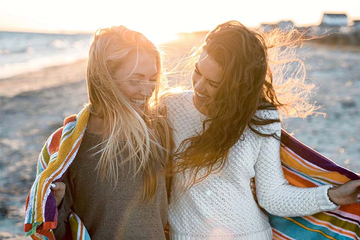 Two women with long hair stand beside each other while holding a blanket on the beach at sunset