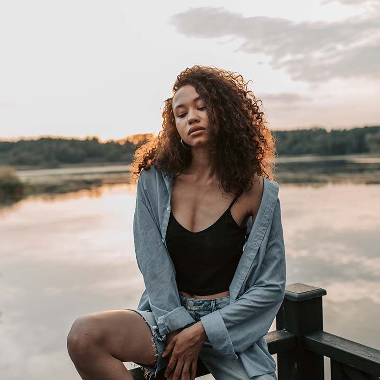 A casually dressed young woman looks forlorn while sitting on a dock with water behind her