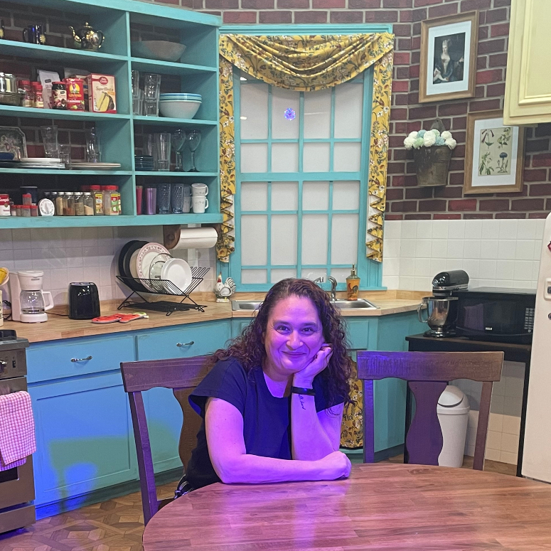 A woman sitting at a wooden dining table with blue cabinetry and shelves and a brick wall behind her