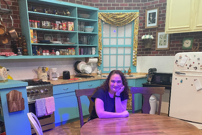 A woman sitting at a wooden dining table with blue cabinetry and shelves and a brick wall behind her