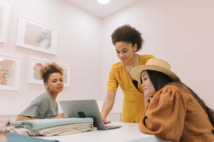 Three women look at a laptop computer that's on a table at work