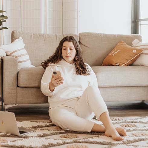 A young woman sits on a cozy rug while looking at her phone