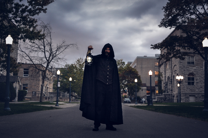 A man in a black hooded cloak holding a lantern on a Kingston street at dusk