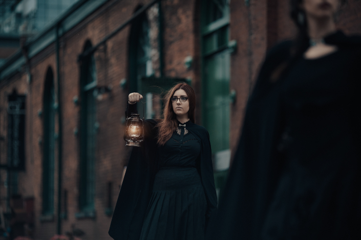 A woman dressed in a black cloak holding a lantern in front of an old distillery building