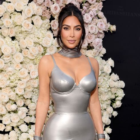 Kim Kardashian poses in front of a floral wall wearing a fitted silver outfit.