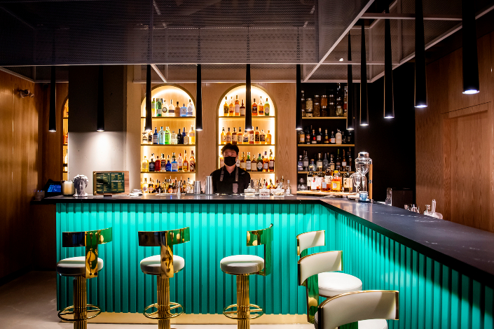 A teal bar with white and gold stools and a bartender standing behind it.