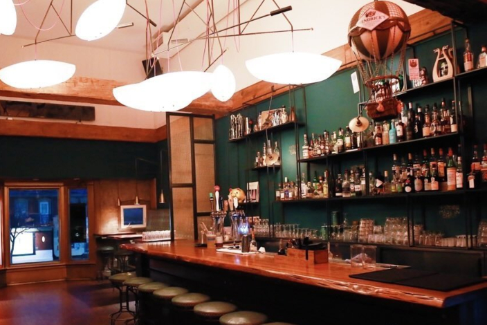 A bar with wooden bartop, stools, open shelves with bottles and large pendant lights
