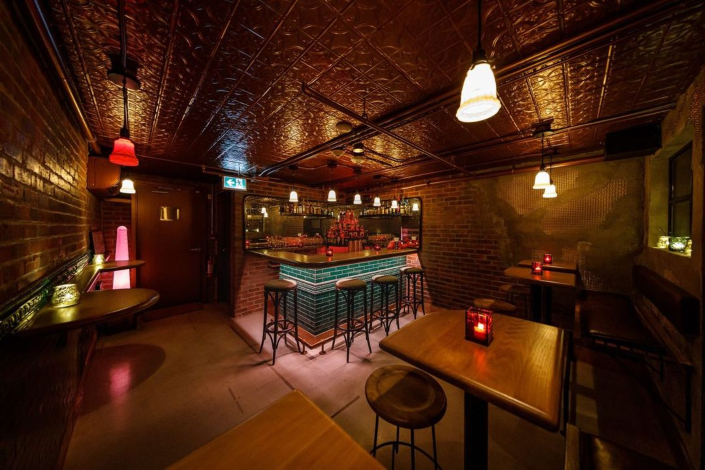 A dimly lit bar with green tile accents, stools, and a wooden table off to the side