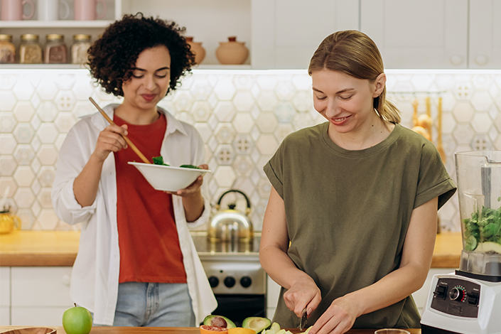 Two young women eating salad and cutting produce in a bright kitchen