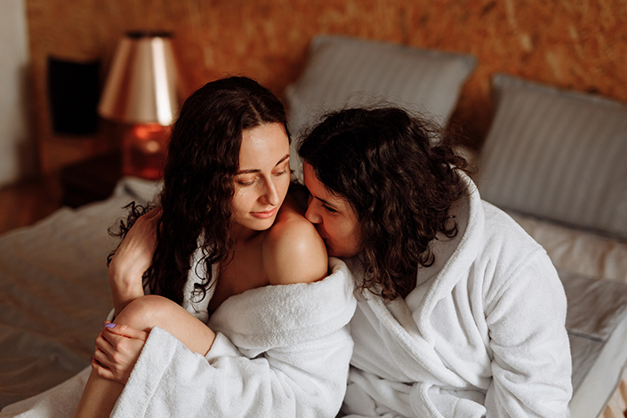 A young couple wearing white robes share an embrace while sitting on a bed
