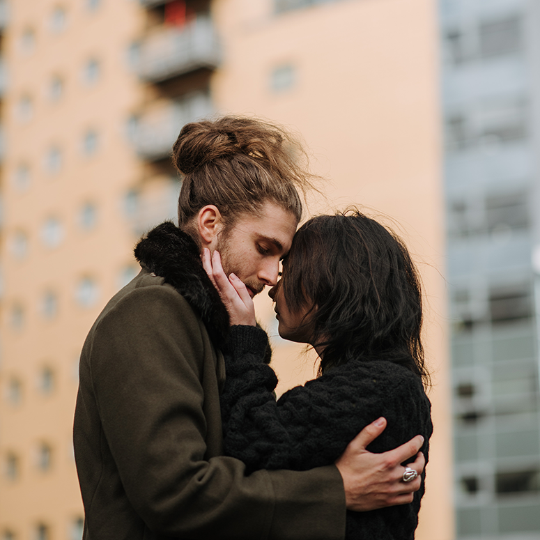 A man with a curly bun and a woman embrace while standing outside