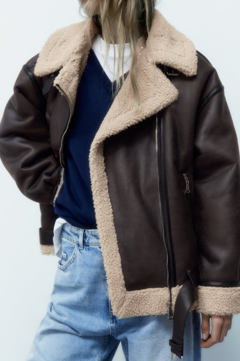 A model wears a brown shearling-trimmed jacket over a blue sweater and jeans