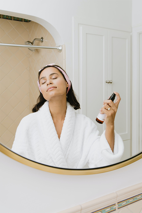 A woman spraying skincare on her face while wearing a white robe