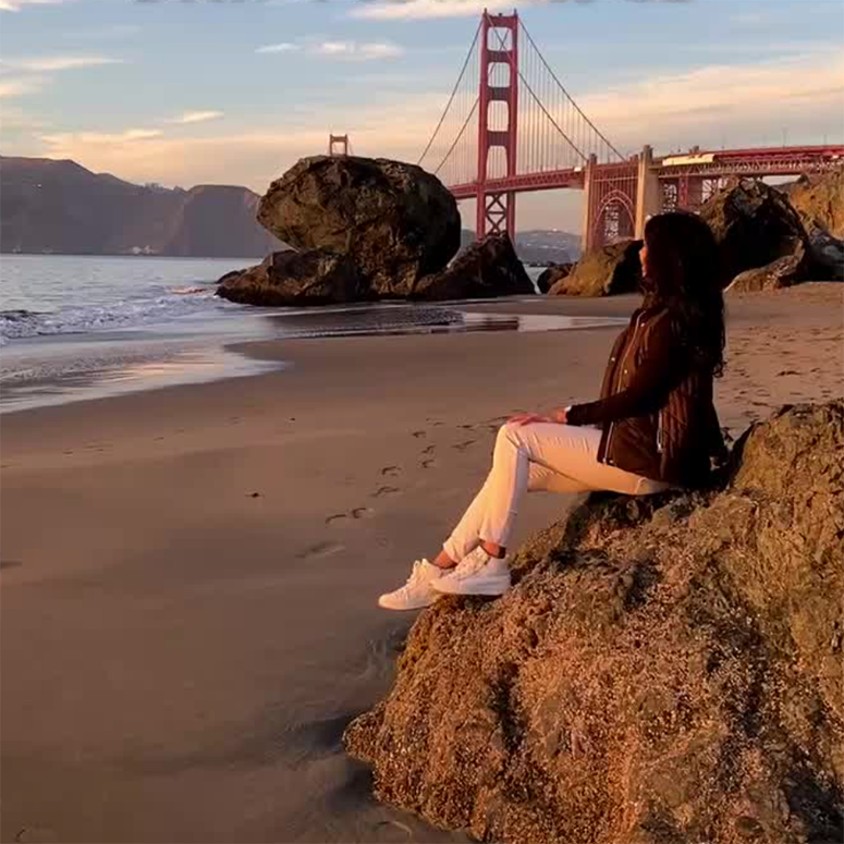 Mari Explores sitting on a rock on a beach looking at the Golden Gate Bridge
