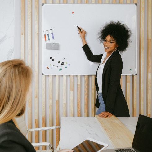 A woman presents at a whiteboard while another woman watches attentively.