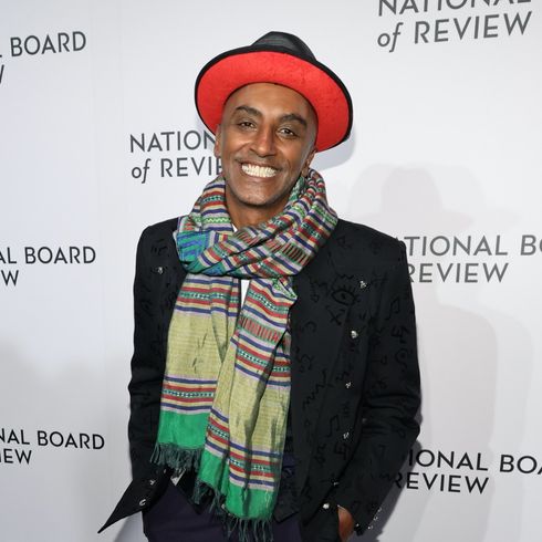 Marcus Samuelsson smiles for the camera on the red carpet wearing a red and black wide-brimmed hat and suit jacket with a chunky multi-coloured scarf.