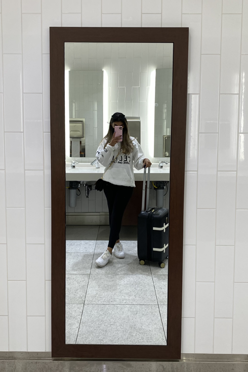 A young woman is casual travel clothes takes a selfie in a mirror at an airport.