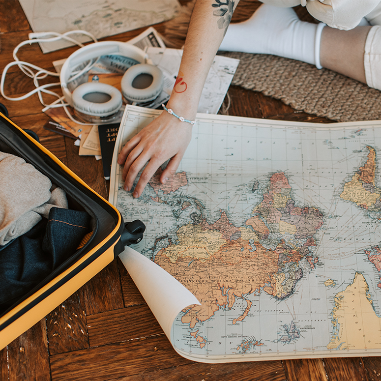 A person puts their hands on a map while sitting on the floor, surrounded by travel gear like a passport and open suitcase