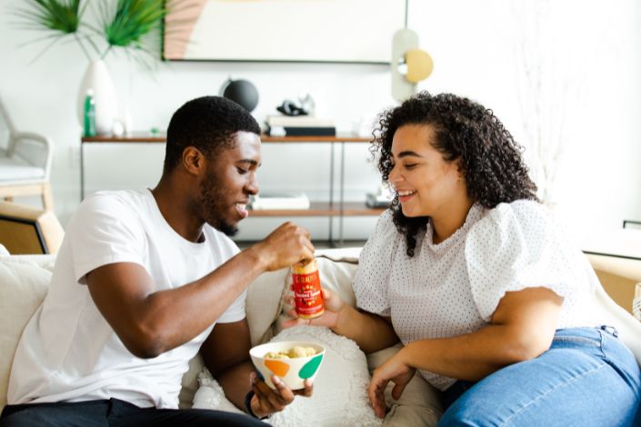 A biracial couples sits together on a couch while sharing a snack.