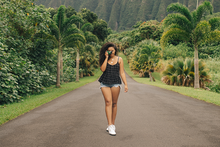 A young woman wearing shorts and a tank top walks down the road, surrounded with lush green trees.