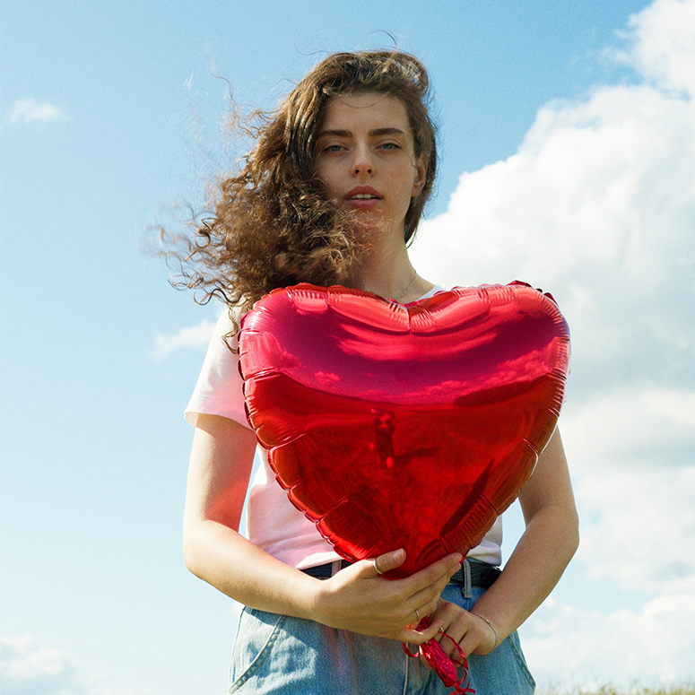 A young woman with curly hair holds a red heart balloon