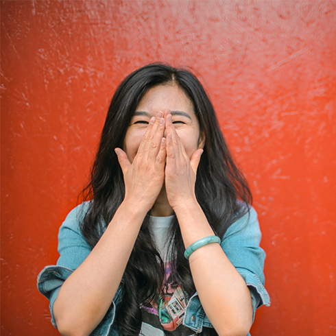 A young woman puts her hands over her face while standing in front of a red wall