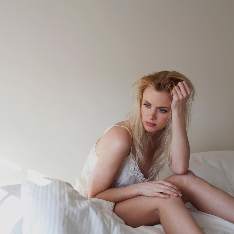 Blonde woman with messy hair sitting on bed with white sheets.