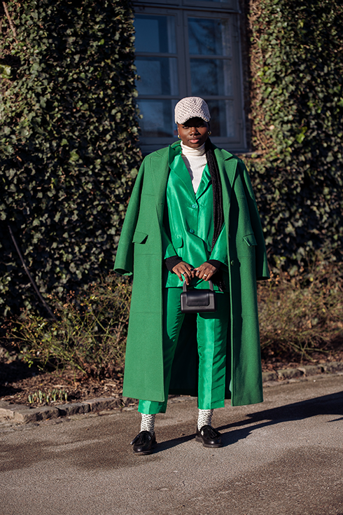 A guest wearing a beige wool cap with Munthe logo print, a white ribbed turtleneck pullover, a green shiny satin blazer, matching green satin suit pants, a green long wool coat, white socks with Munthe logo print, black loafers a black shiny leather handbag outside Munthe during the Copenhagen Fashion Week Autumn/Winter 2023 on February 02, 2023 in Copenhagen, Denmark. 