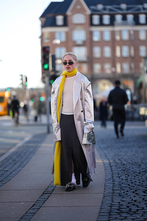 A guest wears black sunglasses from D&G, a silver piercing, a white t-shirt, a white long trench coat, a yellow wool long scarf, a black slit / split long skirt, white socks, silver shiny leather handbag, black shiny leather with brown fur loafers , outside Mark Kenly Domino Tan, during the Copenhagen Fashion Week Autumn/Winter 2023 on February 02, 2023 in Copenhagen, Denmark